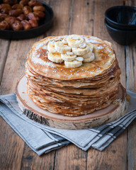 On the table on a stand made of wood cut a pile of thin pancakes, sprinkled with jam, sprinkled with powdered sugar, on top are banana and peanuts. In the background a plate with dates. Style Rustic