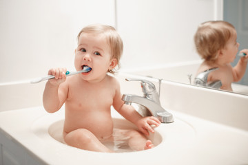 Portrait of cute Caucasian blonde funny baby girl boy sitting in washroom sink with water. Child...
