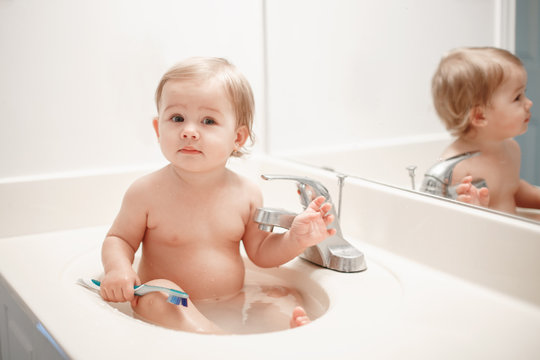 Portrait of cute Caucasian blonde funny baby girl boy sitting in washroom sink with water. Child brushing teeth. Happy healthy childhood concept. Everyday home family sweet moment