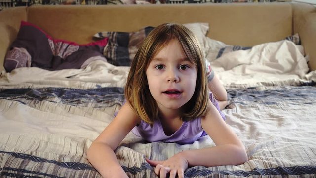 A girl with blonde hair and pigtails, of European or American appearance, is lying on the bed. To the child of 5-6 years. She is watching a movie. Close-up portrait.
