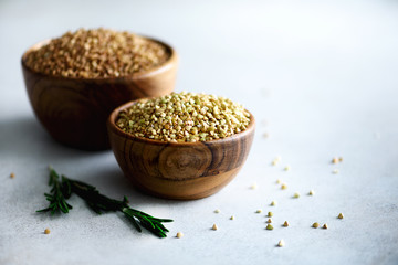 Raw organic green and roasted buckwheat in wooden bowls and rosemary on grey background. Copy space. Food ingredients.