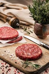 On a wooden cutting board on kraft paper there are raw beef burgers for burgers, spices, rosemary, a pepper mill, a cooking process. Top view