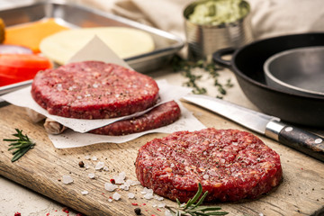On a wooden cutting board on kraft paper there are raw beef burgers for burgers, spices, rosemary, a pepper mill, a cooking process. Top view