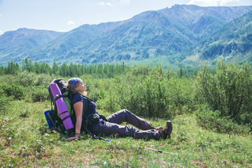 Naklejka na ściany i meble Woman hiker with backpack sitting on green grass and feel relax with closed eyes at background of high mountains