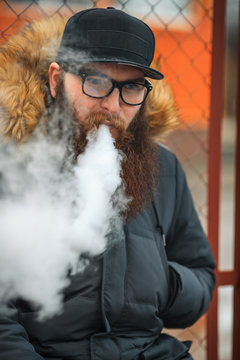 Vape bearded man in real life. Portrait of young guy with large beard in glasses and a cap vaping an electronic cigarette and letting steam opposite the fence in the autumn.
