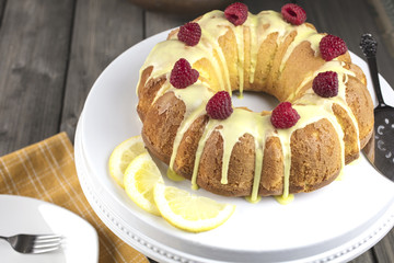 Lemon cake frosted with yellow sugar icing and red raspberries, dressed with lemon slices on the side on a white platter