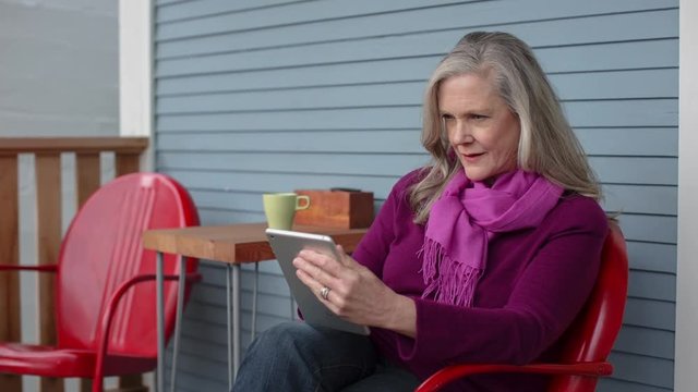 Handheld shot of senior woman using tablet computer while sitting on chair at porch