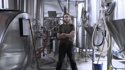 Dolly shot of confident brewer standing arms crossed amidst storage tanks in brewery