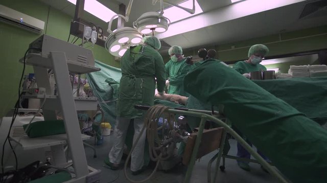 Surgeon team of doctors and medical assistant operating hip of patient under anesthesia on table, surgery procedure at operation room, surgical tools and instruments on tray, crane shot, tilt up