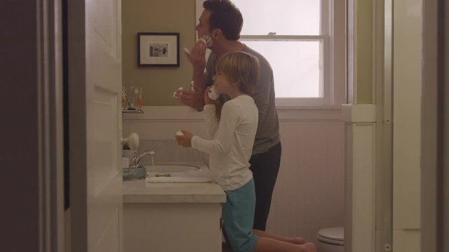 Handheld shot of father with son applying shaving cream in bathroom seen through doorway