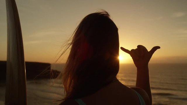 SLOW MOTION CLOSE UP: Unrecognizable girl with long flowing hair gazing at golden sunset giving the shaka sign. Female surfer making hang loose signal with her hand. Woman using Hawaiian hand gesture