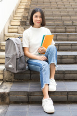 Portrait of Attractive Asian Student Woman sitting at outdoor place. Woman holding Book with Attractive Smiling to Camera. People with Education Concept.