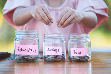 Cute asian little child girl putting coin into glass bottle in the garden. Kid saving money for the...