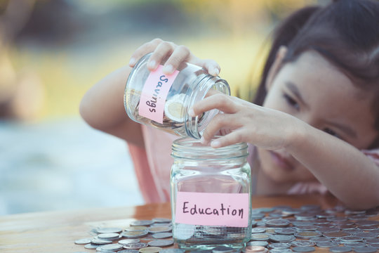 Cute Asian Little Child Girl Putting Coin Into Glass Bottle In The Garden. Kid Saving Money For The Future Concept