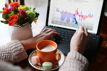 Woman working on a laptop and drinking coffee. Working space with cup of coffee on a foreground in the focus between hands of woman, analyzing data on computer in shallow depth of field.
