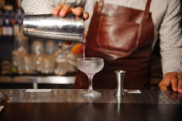 barman in a brown apron pours from a steel shaker alcohol cocktail