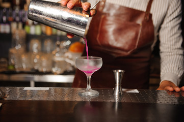 bartender pours from a steel shaker into a cocktail glass a lilac-colored alcohol cocktail