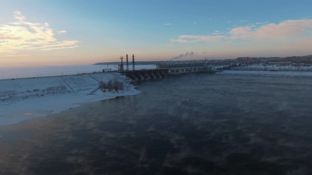 Fly over soaring ice water and approaching the building of a power station. Novosibirsk reservoir and hydroelectric power station: frozen water and sun, reflected in the ice crust. Aerial view.