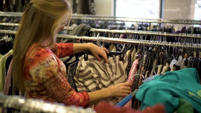 Closeup tracking steadicam shot of woman shopping and flipping clothes on a rack in a second hand clothing store.