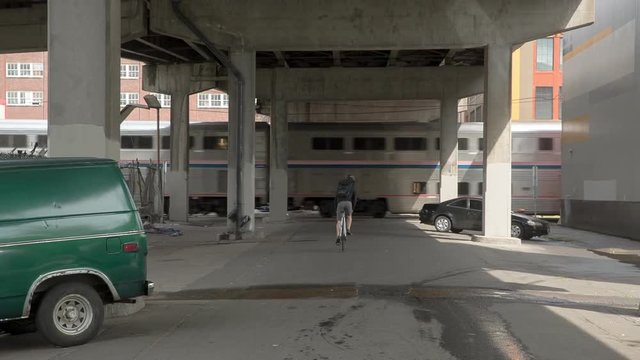Handheld shot of male commuter riding bicycle leading towards moving train under bridge