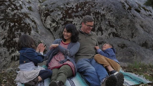 Slow motion dolly shot of cheerful family sitting on picnic blanket by rock in forest