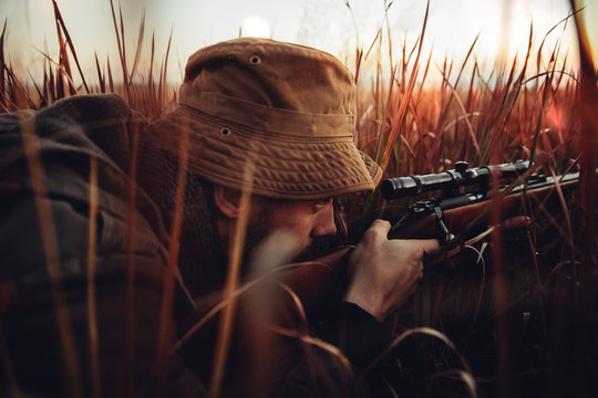 Hunter laying in long red grass looking trough his sniper