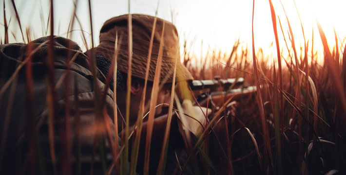 Hunter with rifle laying in long grass and concentrating on shooting prey