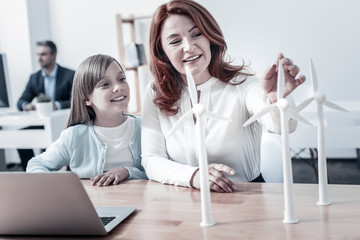Taking care of nature. Cheerful mother and daughter smiling while spending day together and talking about a set of wind turbines decorating a table at work. © zinkevych