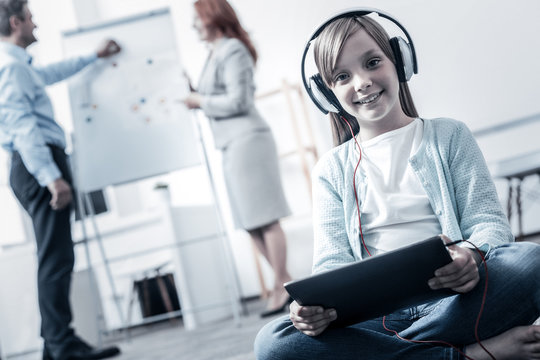 Favorite song. Low angle shot of a cute little girl sitting on the floor with a tablet computer in her hands while listening to mucis and waiting for her parents working in the background.