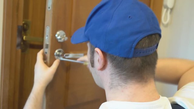 Young handyman in uniform fixing door lock.