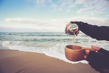 Man traveler hands poured from a thermos bottle cup of coffee, sitting near the fire in autumn forest.