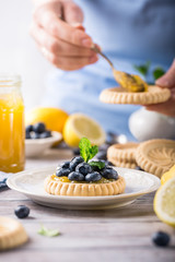 Shortbread homemade tartlet filled with lemon curd and fresh blueberries with women's hands on rustic wooden background. Holiday food concept.