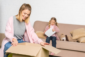 Fototapeta na wymiar smiling woman packing cardboard box while little daughter using digital tablet