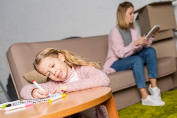 cute little child drawing at table while mother using digital tablet on sofa