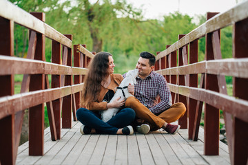 Young stylish happy couple in love kissing with little white dog on wooden bridge. Pretty girl and man having fun with maltese outdoors.