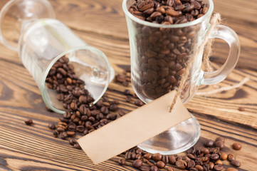 Lot of fried coffee beans in transparent glass for mulled wine with rectangle blank paper on rope near coffee beans poured out of glass on old wooden rustic brown planks