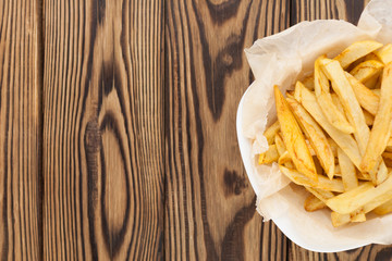Tasty sticks of french fries in white ceramic bowl with crumpled paper on old brown rustic wooden table