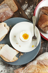 boiled egg on a plate, freshly baked bread and sandwiches with bacon on a wooden table