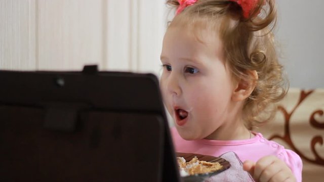 A little cute girl eats muesli and watches cartoons on the tablet. Child and gadgets, applications, internet, games.
