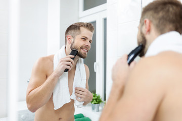 Handsome bearded man trimming his beard with a trimmer - obrazy, fototapety, plakaty