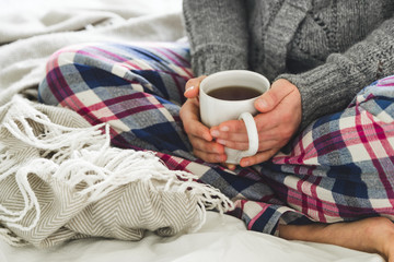 Young woman in cozy pyjamas and gray cardigan sitting on a bed with a mug of tea
