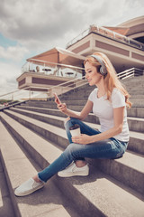 Vertical profile side view photo of attractive cheerful careless stylish wearing casual jeans white tshirt blonde woman enjoying the sound in her earphones, using mobile and drinking takeaway tea