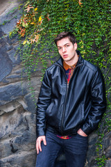 American man autumn/winter casual street fashion. Young handsome man wearing black leather jacket, pants, standing by rock wall with long leaves at Central Park, New York, looking at you...