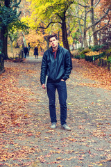 Young American man traveling at Central Park, New York in autumn day. Man wearing black leather jacket, jeans, gray casual shoes, holding laptop computer, walking on road with colorful trees, leaves