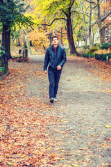 Young American man traveling at Central Park, New York in autumn day. Man wearing black leather jacket, jeans, gray casual shoes, holding laptop computer, walking on road with colorful trees, leaves