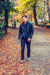 Young American man traveling at Central Park, New York in autumn day. Man wearing black leather jacket, jeans, gray casual shoes, holding laptop computer, walking on road with colorful trees, leaves