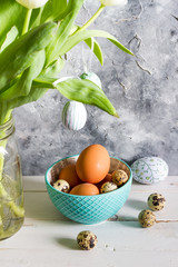 Easter eggs in blue bowl. White tulips in the jar. Gray concrete background, white wooden table. 