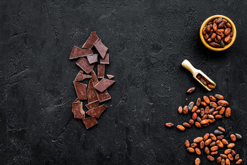 Chocolate and cacao concept. Cocoa powder in bowl near cocoa beans and broken chocolate on black background top view copy space