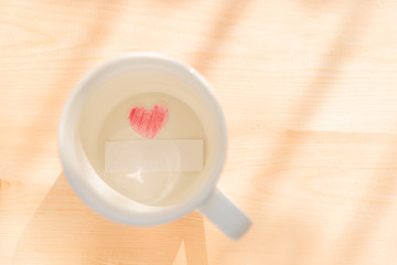 White coffee cup with paper cut in heart shape painted with red pencil and empty or blank paper card on wood background under sunlight to show love relationship on Valentines day or wedding party