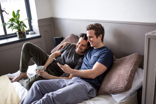 Young male couple sitting on bed and looking at smartphone together